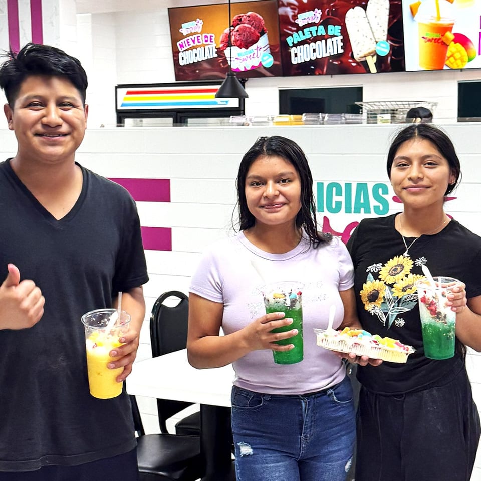 Three smiling people hold colorful drinks and fruit desserts in a bright, modern ice cream shop.