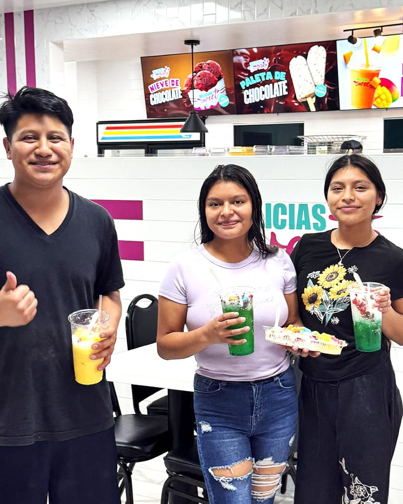 Three smiling people hold colorful drinks and fruit desserts in a bright, modern ice cream shop.