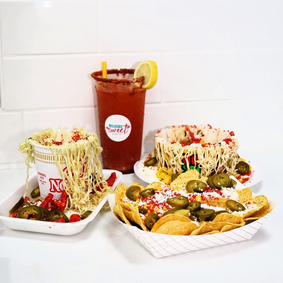 A red drink with lemon, a cup of noodles, and nachos with jalapeños on a white surface.