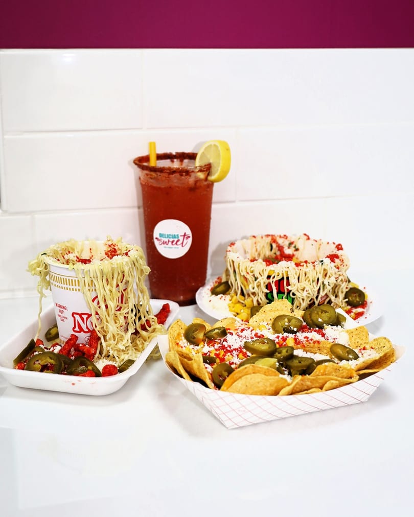 A red drink with lemon, a cup of noodles, and nachos with jalapeños on a white surface.