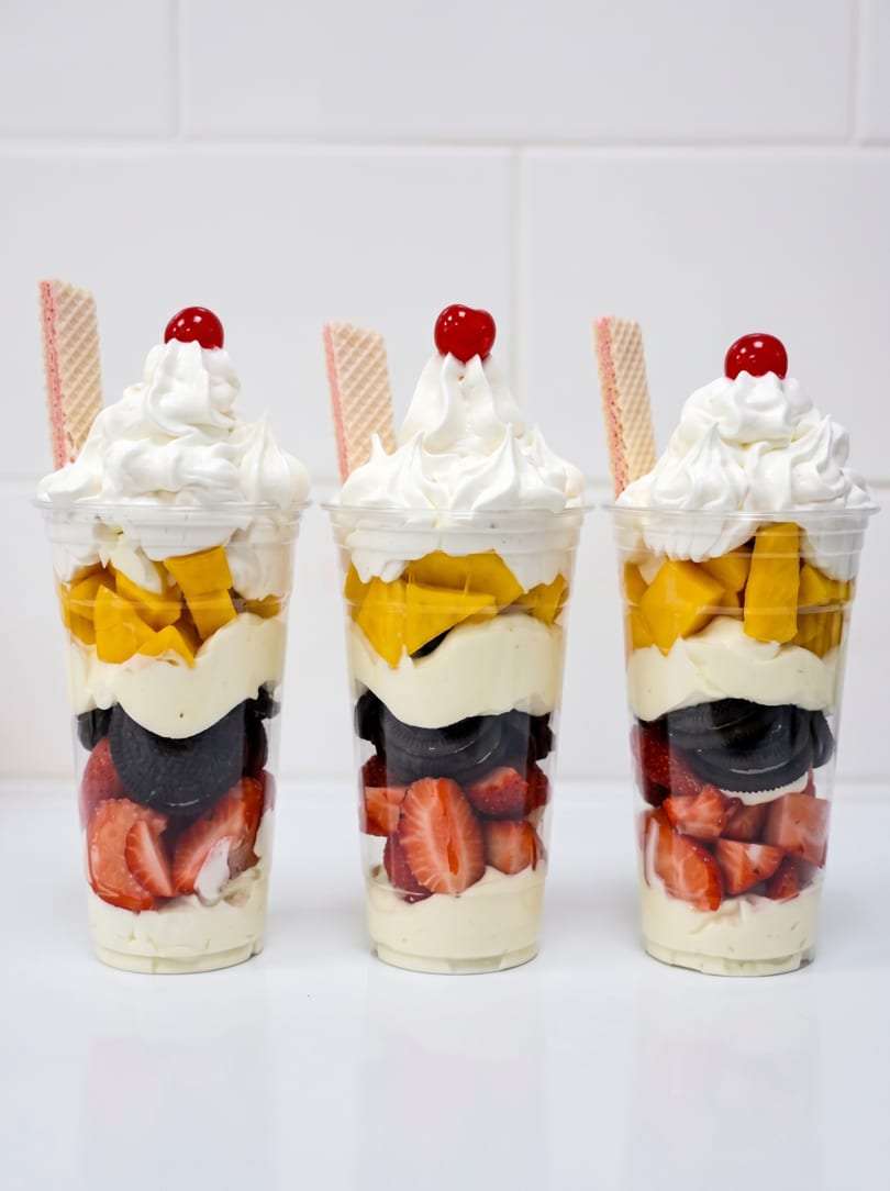 Three parfaits with layers of strawberries, cookies, cream, mangoes, whipped cream, and cherries.