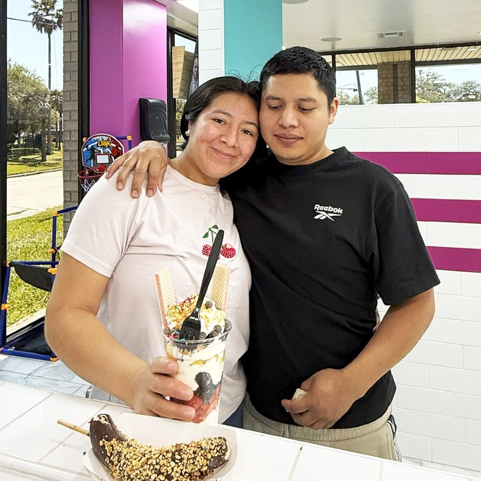 A smiling couple stands behind a counter with a large parfait and a chocolate-covered banana.