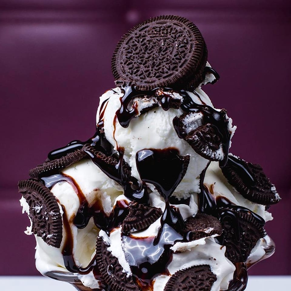 Vanilla ice cream sundae with chocolate syrup and Oreo cookies against a purple background.