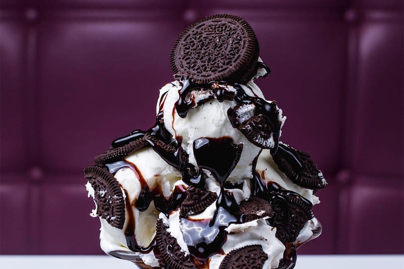 Vanilla ice cream sundae with chocolate syrup and Oreo cookies against a purple background.