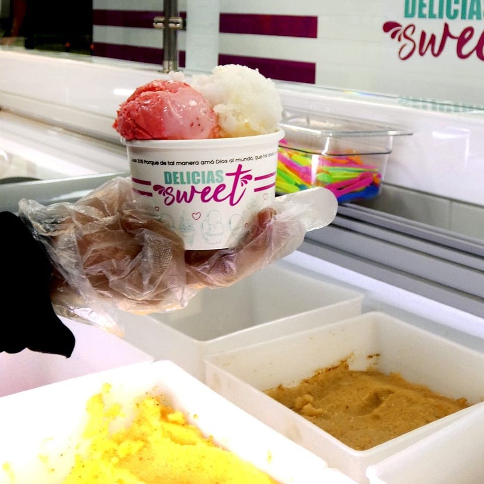 A gloved hand holds a cup of Delicias Sweet ice cream with strawberry and coconut scoops.
