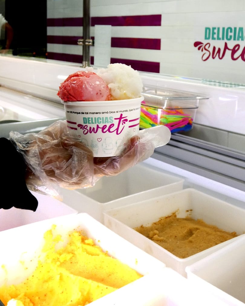 A gloved hand holds a cup of Delicias Sweet ice cream with strawberry and coconut scoops.