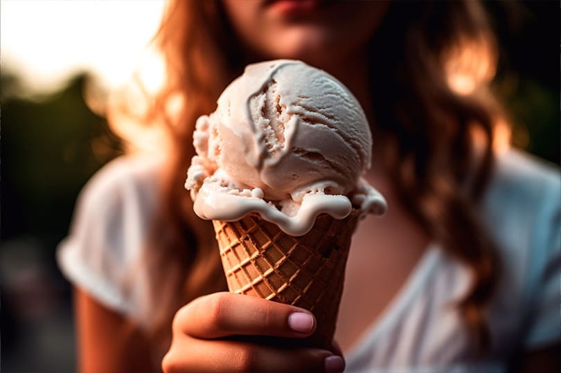 A person holds a waffle cone with two scoops of melting vanilla ice cream at sunset.