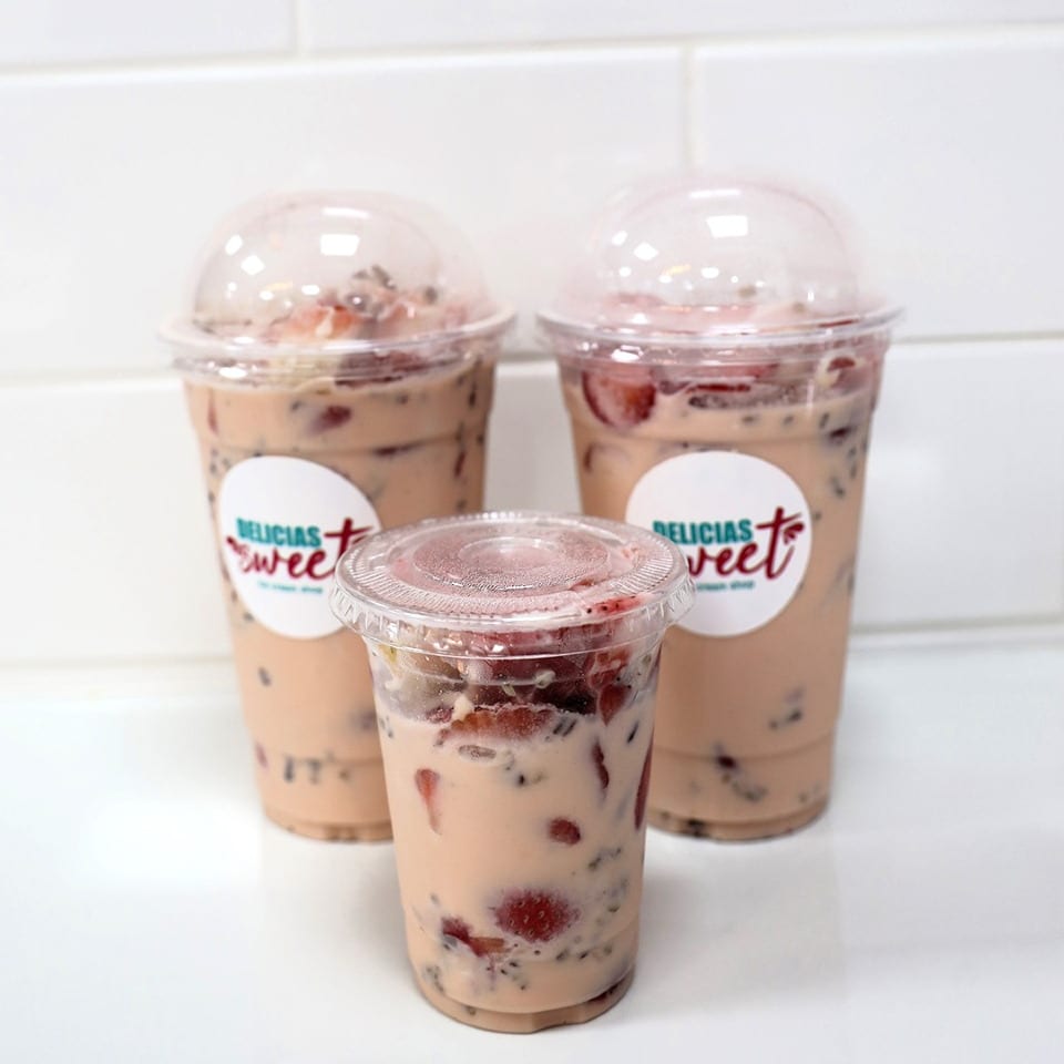 Three Delicias Sweet strawberry milk drinks with chia seeds and fresh strawberries on a white counter.