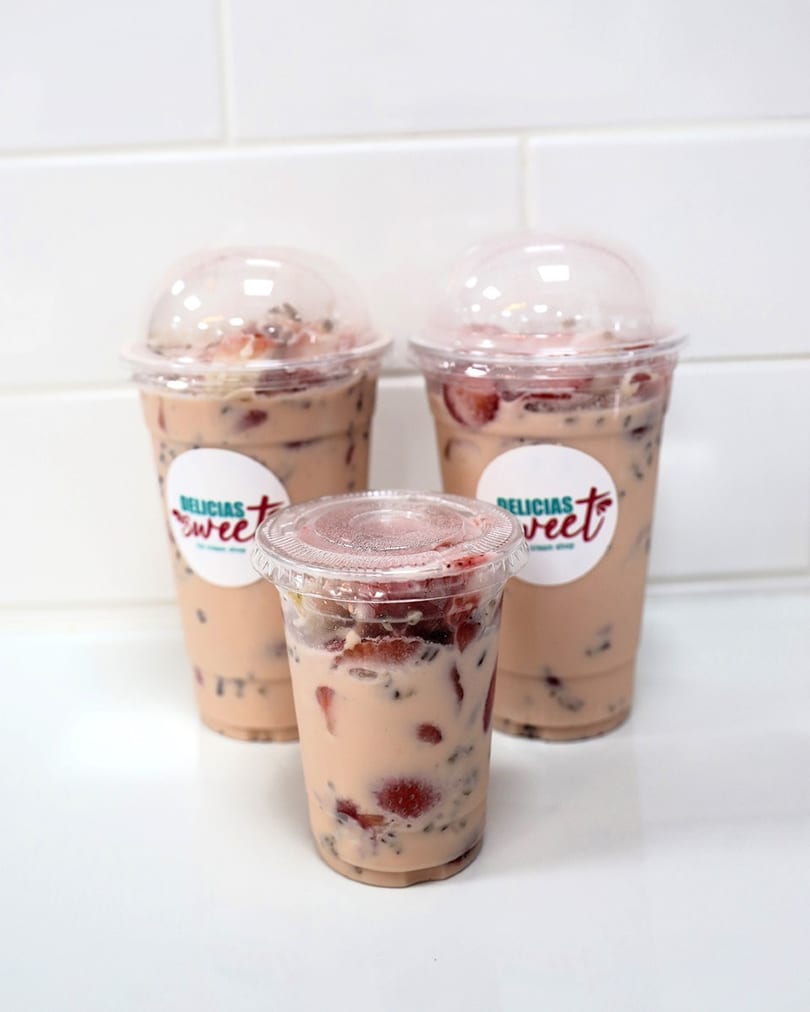 Three Delicias Sweet strawberry milk drinks with chia seeds and fresh strawberries on a white counter.