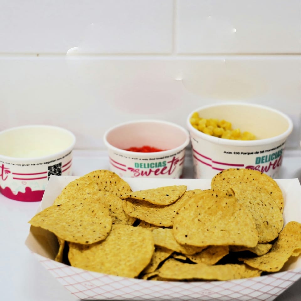 Nachos in a paper tray with three small cups of toppings, including corn and red sauce.