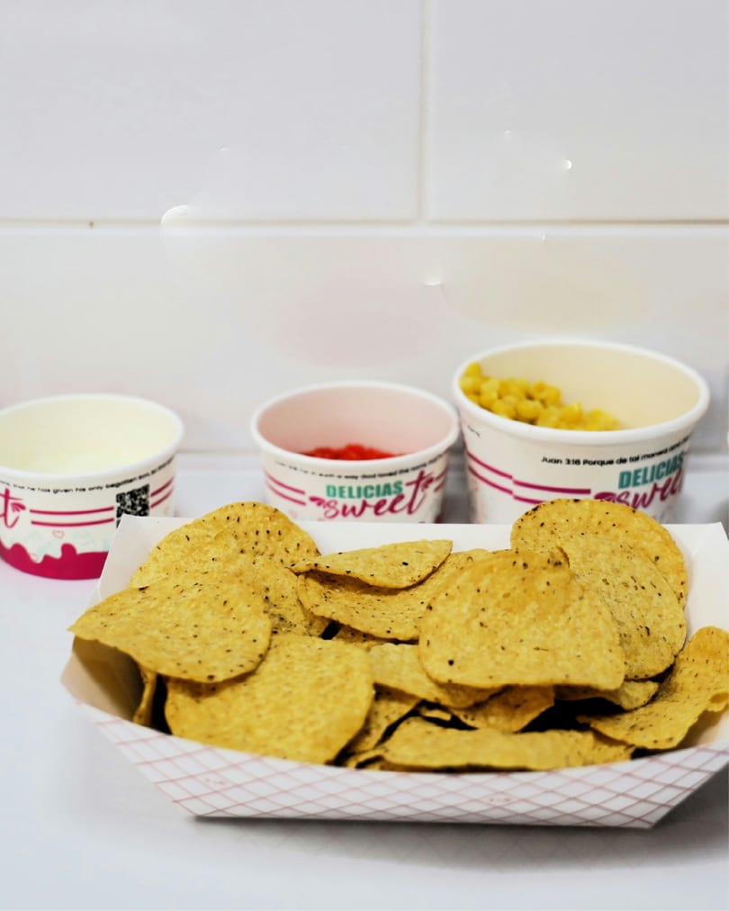 Nachos in a paper tray with three small cups of toppings, including corn and red sauce.