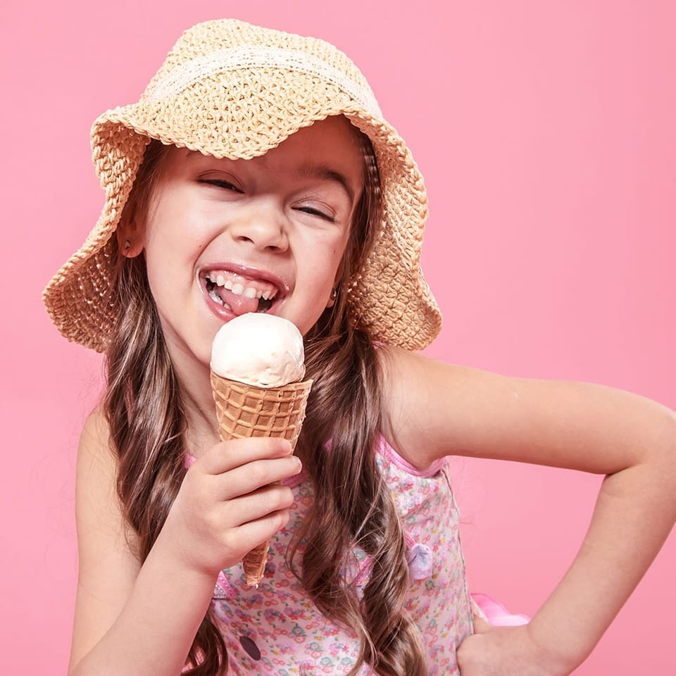 A happy young girl in a straw hat and floral top licks a vanilla ice cream cone.