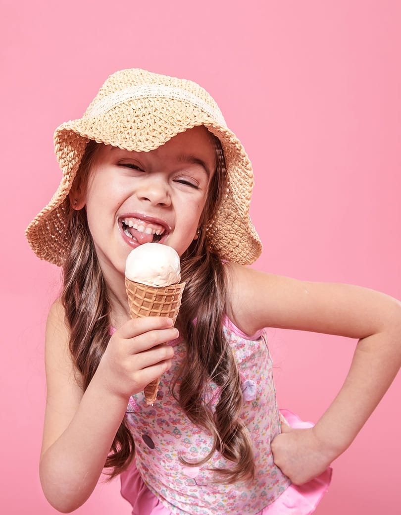 A happy young girl in a straw hat and floral top licks a vanilla ice cream cone.