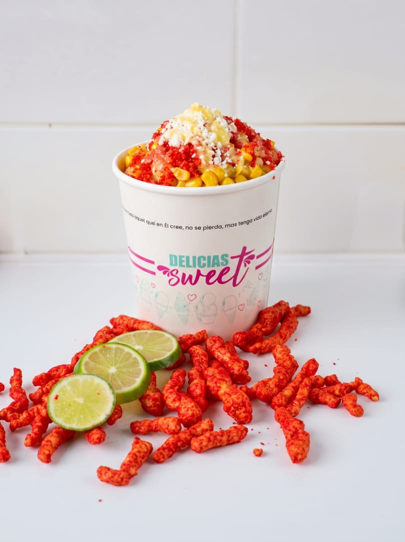 A cup of esquites with cheese, chili, and corn, surrounded by lime slices and spicy snacks.