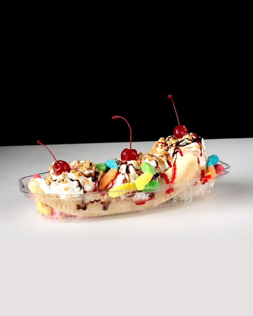 A banana split in a clear plastic dish, topped with whipped cream, cherries, nuts, and gummy candies.