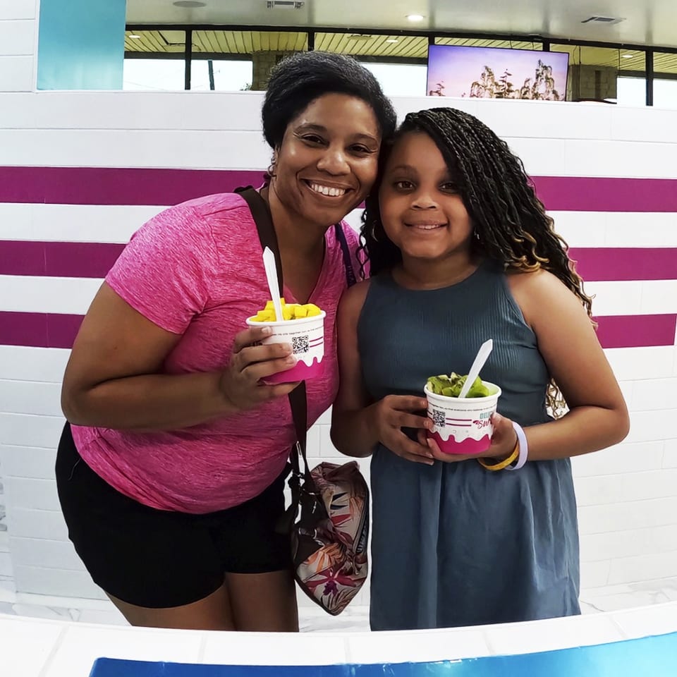 A smiling woman and girl hold cups of frozen yogurt with fruit toppings in a brightly lit shop.