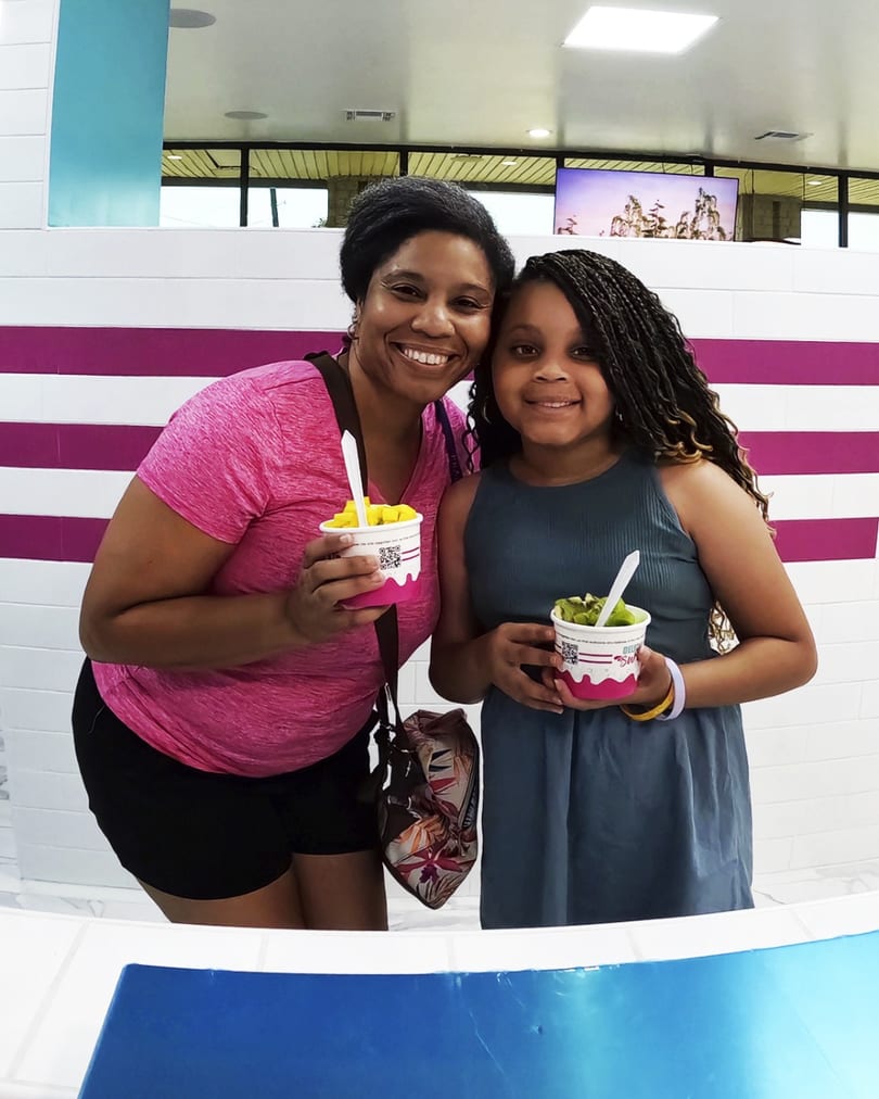 A smiling woman and girl hold cups of frozen yogurt with fruit toppings in a brightly lit shop.