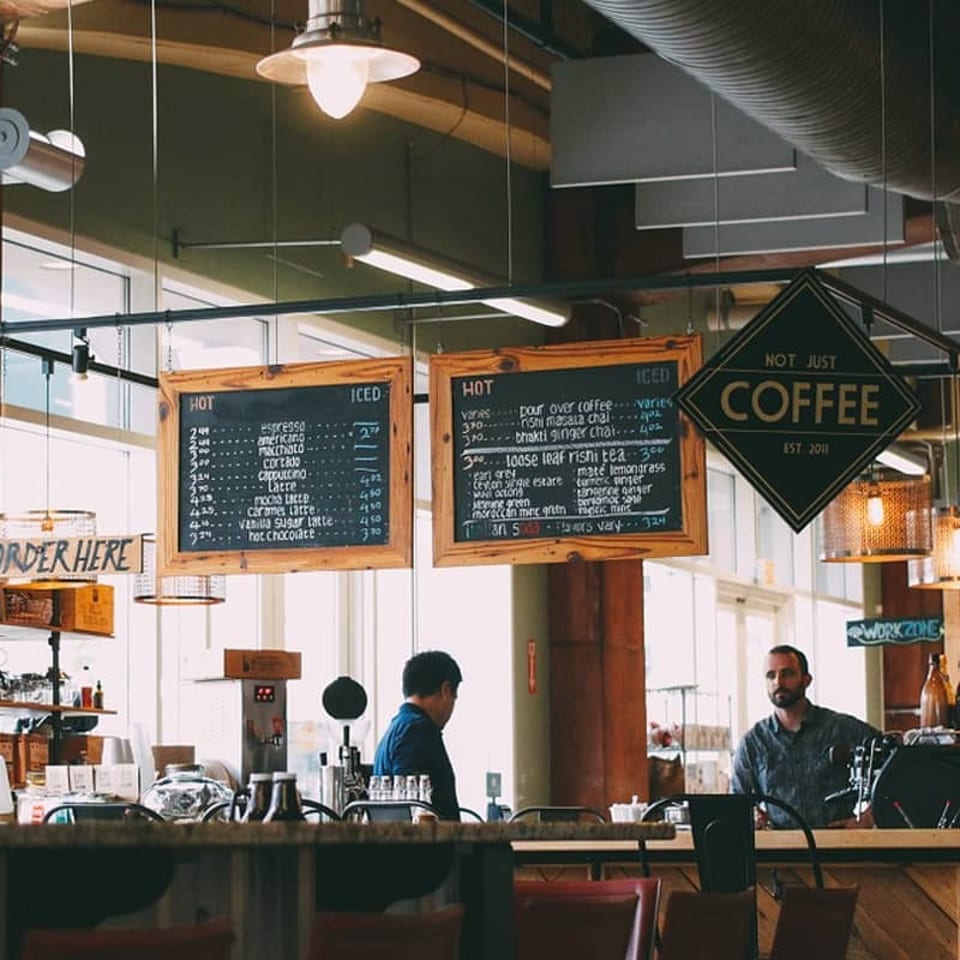 Two men stand behind a coffee shop counter with menus and a "Not Just Coffee" sign hanging above.